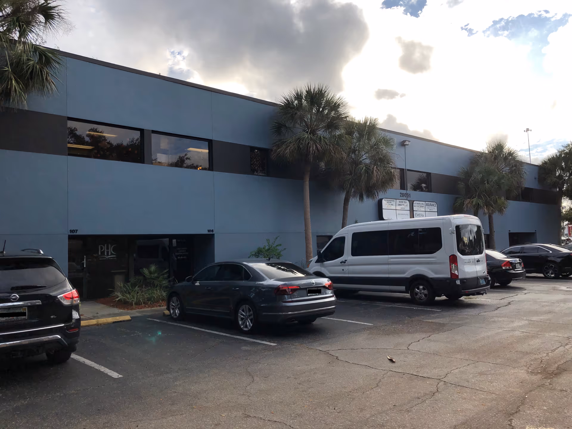Blue-gray facility exterior with palm trees and a parking lot in front.