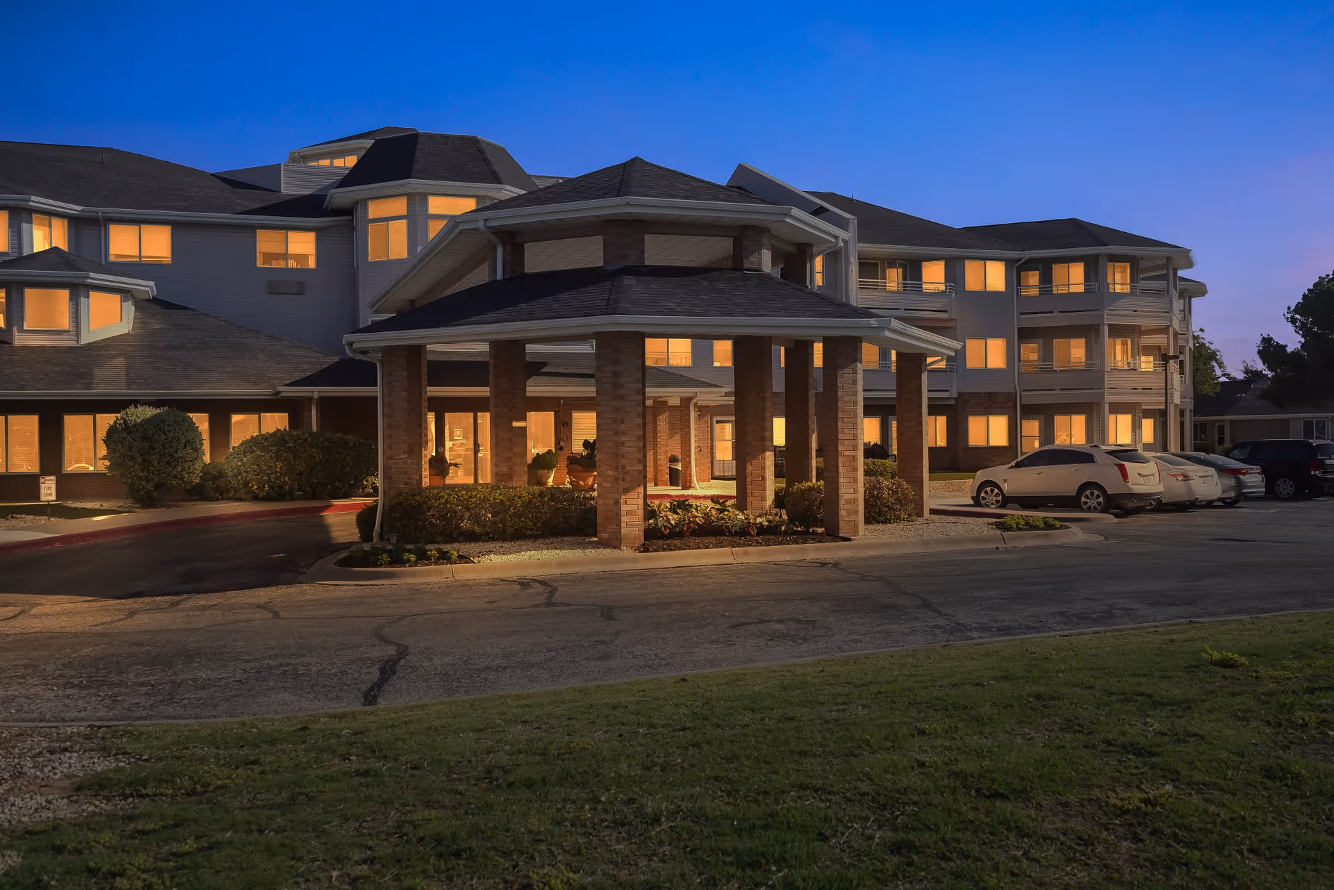 Front entrance of a multi-story senior living building at dusk with a covered porte-cochere, illuminated windows, and parked cars.