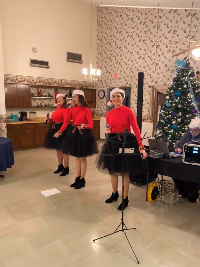 Three women dressed in red tops, black skirts, black ankle boots, and Santa hats are singing indoors. They are holding microphones and standing in front of a decorated Christmas tree. To the right, a person is seated at a table with audio equipment and a laptop. The room has floral wallpaper and wooden cabinets in the background.