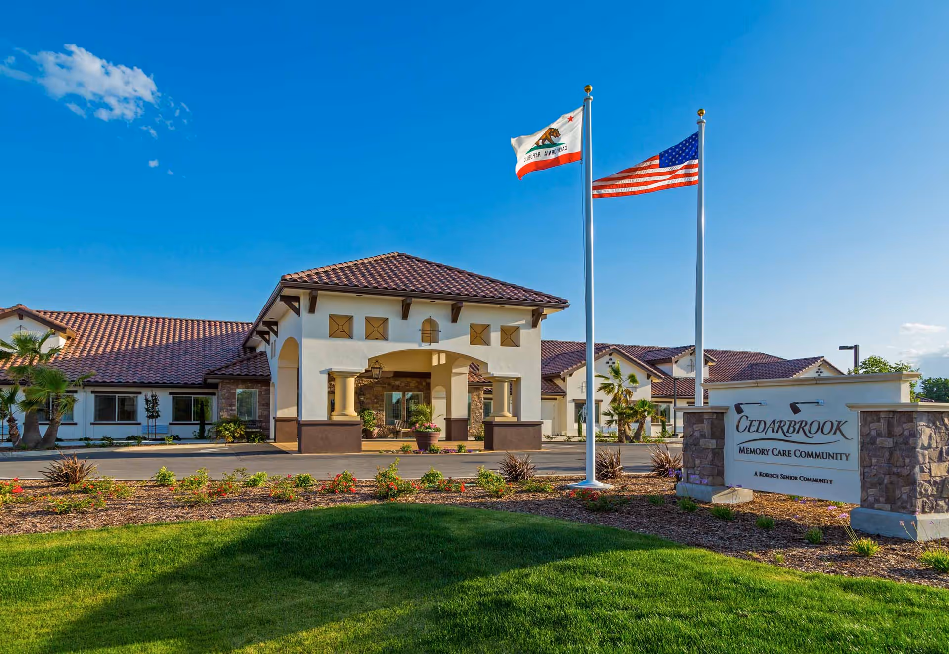 Exterior view of Cedarbrook Memory Care Community building with a covered entrance, two flagpoles displaying the California state flag and the American flag, landscaped garden with green grass and plants, and a clear blue sky.