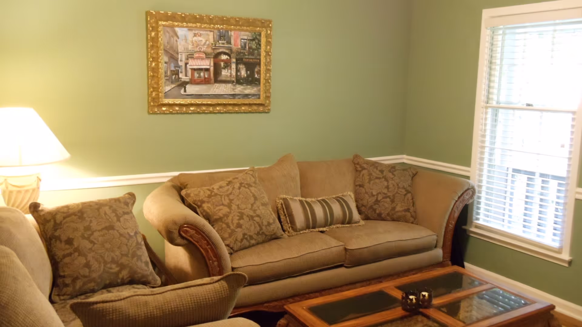 A cozy living room with a beige sofa adorned with patterned cushions and a striped lumbar pillow. A wooden coffee table with a glass top sits in front of the sofa. The walls are painted green with white trim, and a framed painting of a street scene hangs above the sofa. A window with white blinds allows natural light into the room, and a table lamp with a beige shade is visible on the left side.