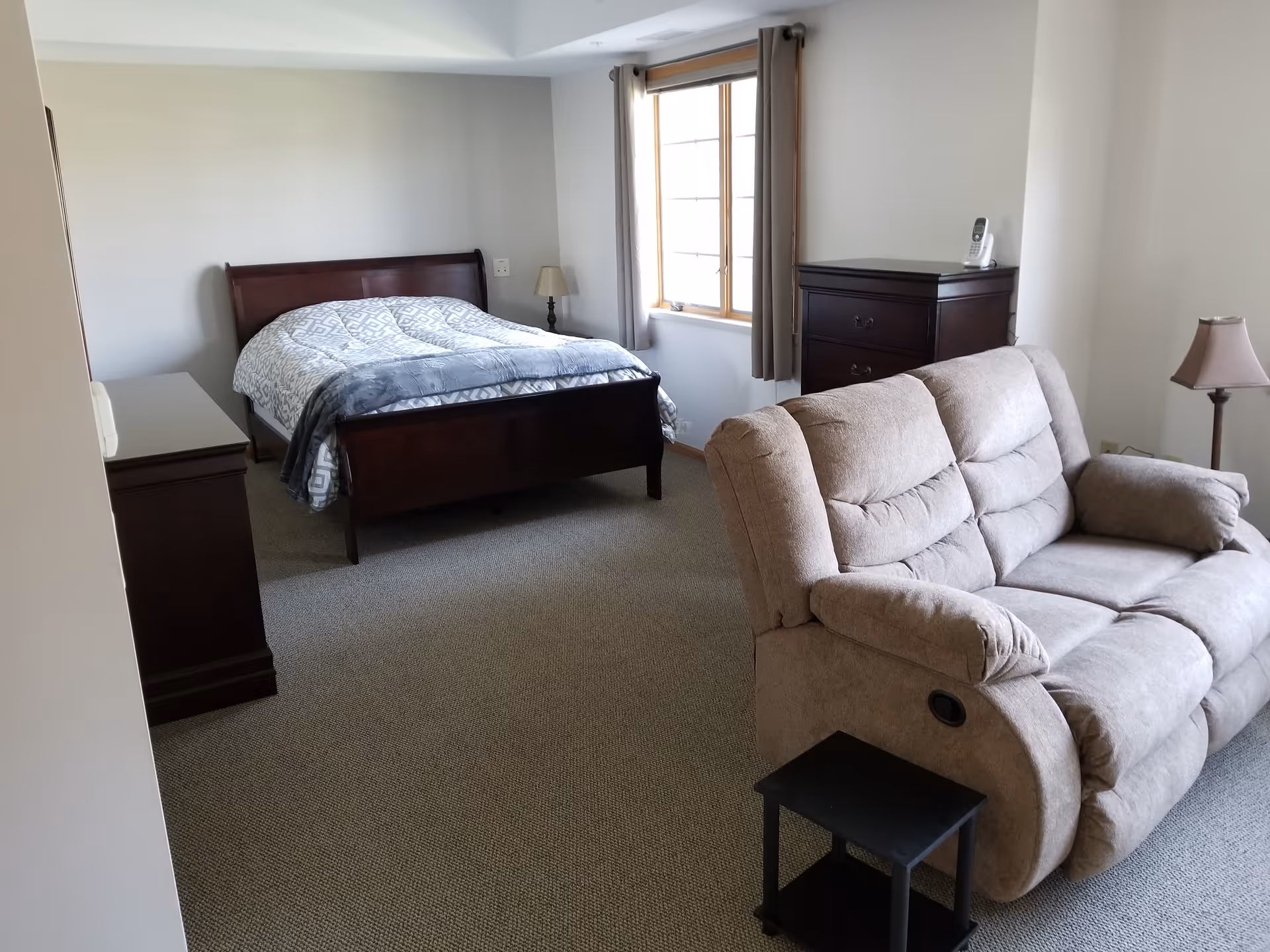A furnished senior living bedroom with bed, sofa, dresser, and window.