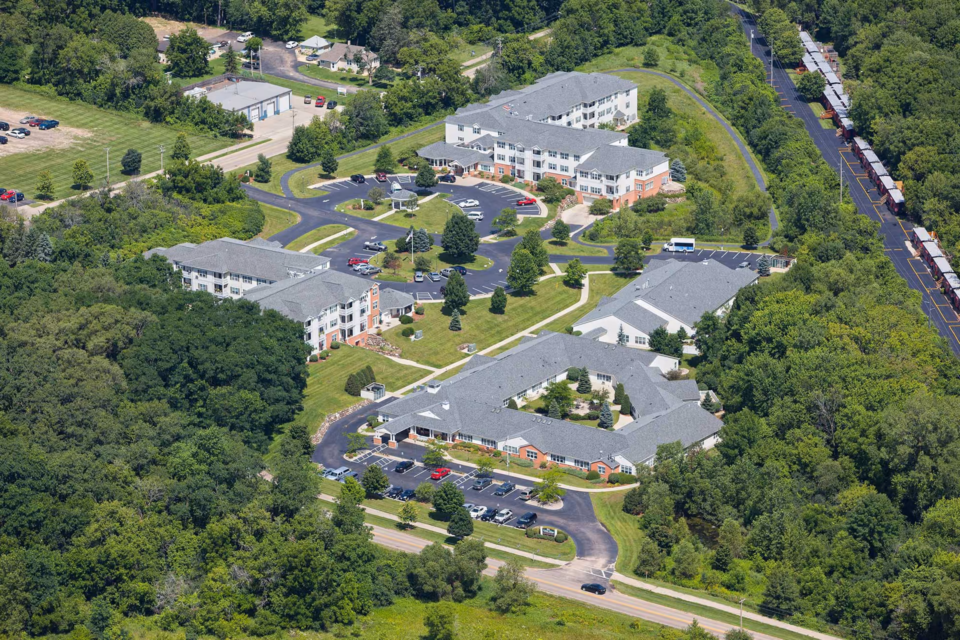 Aerial view of a memory-care campus showing several multi-wing buildings, parking lots, pathways, and surrounding trees.