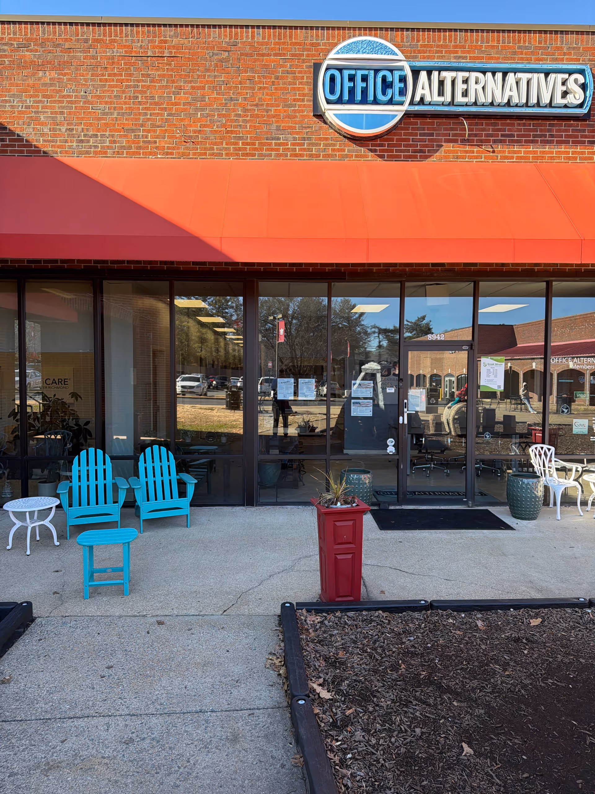 Brick storefront with a red awning and blue chairs outside a glass entrance.