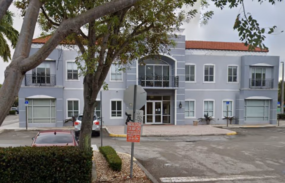 Gray two-story building exterior with entrance, parked cars, and tree-lined frontage.