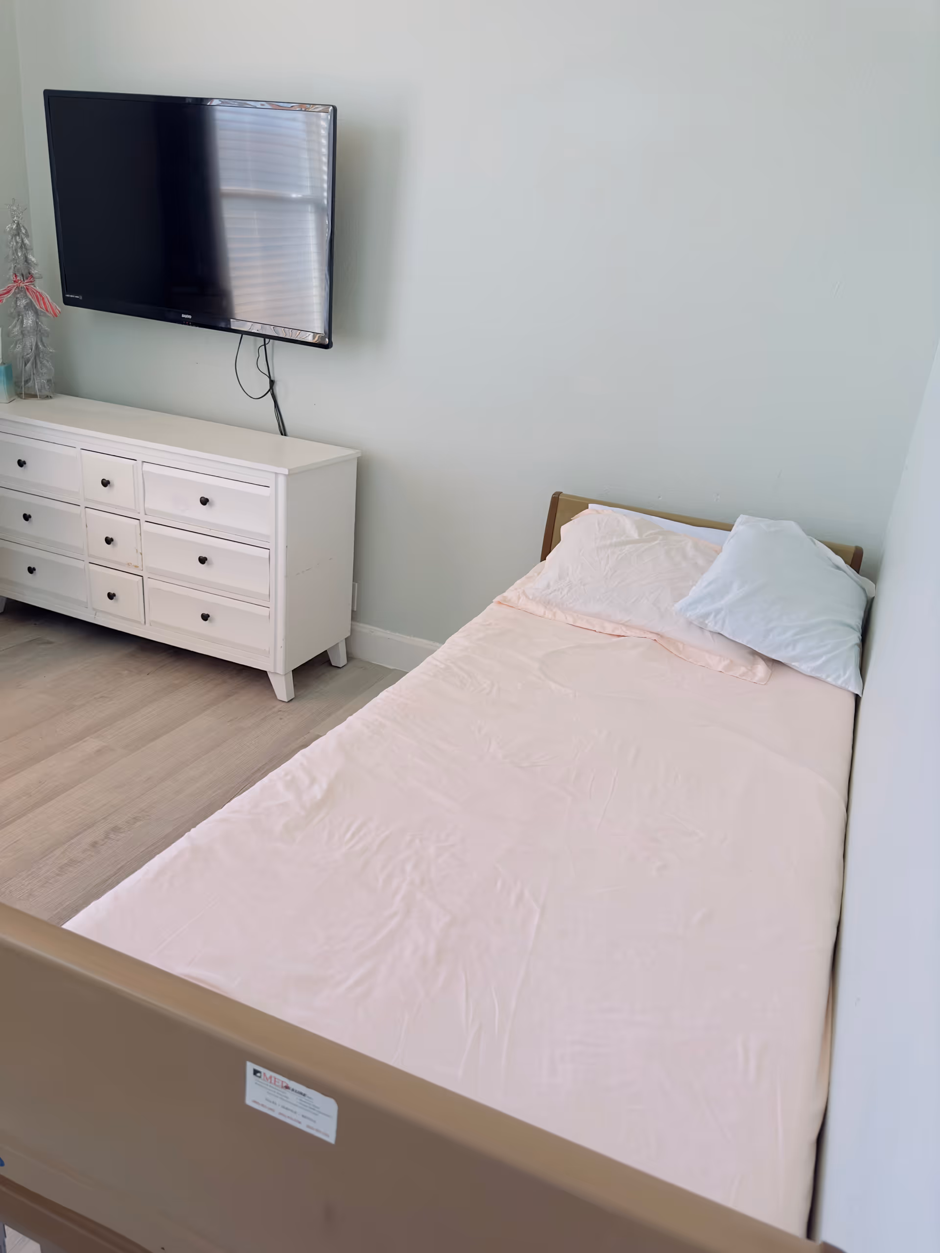 A bedroom with a wall-mounted TV, white dresser, and a pink-sheeted bed.