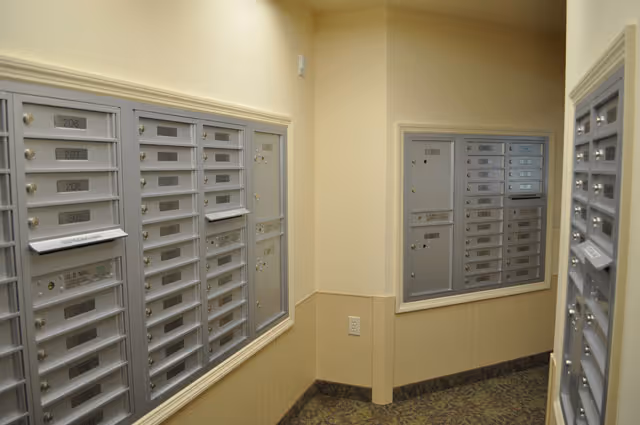 Indoor mailroom area with multiple silver mailboxes mounted on beige walls. The mailboxes are labeled with numbers and some have mail inside. The floor has patterned carpet and the walls are painted in a light beige color.