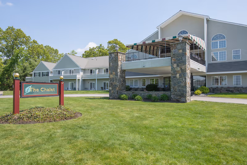 Exterior view of a senior living facility named The Chalet, featuring a large building with multiple windows, a covered outdoor balcony supported by stone pillars, and a well-maintained green lawn with a sign displaying the facility's name.
