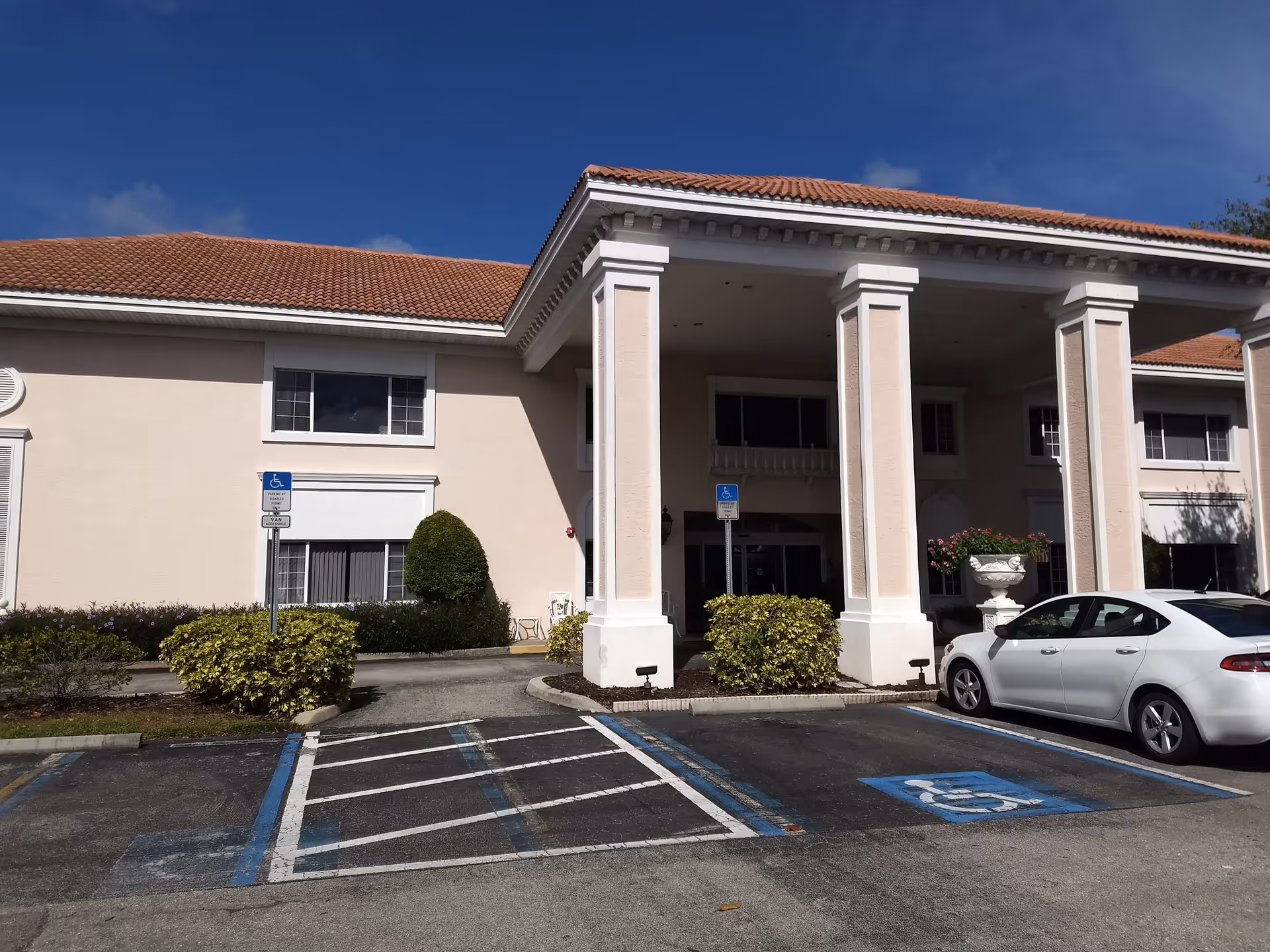 Exterior view of a senior living facility building with a covered entrance supported by large white columns. There are handicap parking spaces in front of the entrance, one of which is occupied by a white car. The building has a beige facade and a red-tiled roof under a clear blue sky.