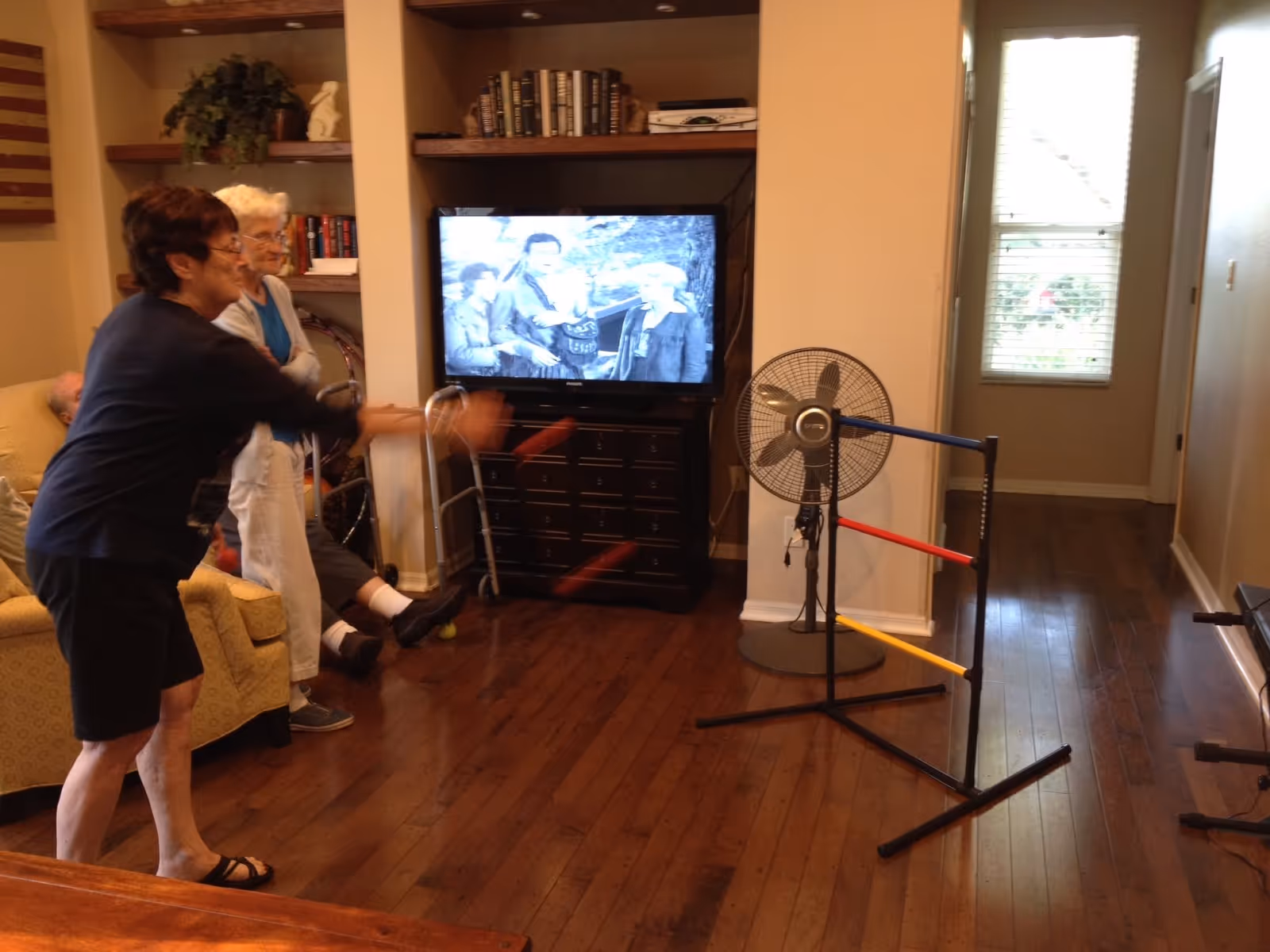 Senior residents play a ring-toss game in a living room with a TV, shelves, and a standing fan.