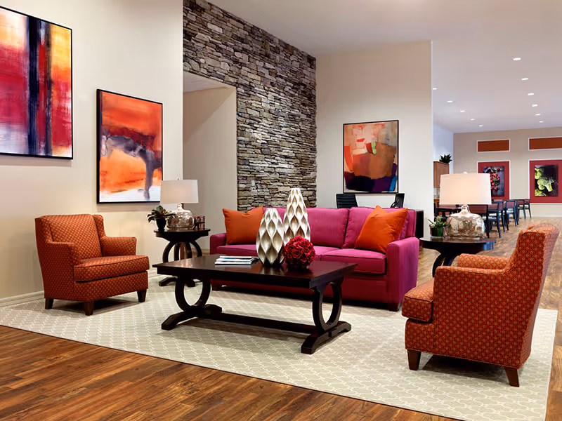 A modern living room area with a magenta sofa adorned with orange pillows, two orange patterned armchairs, a dark wooden coffee table with decorative vases and a flower arrangement, and side tables with lamps. The room features a stone accent wall and colorful abstract paintings on the walls. The floor is wooden with a light patterned rug under the furniture.
