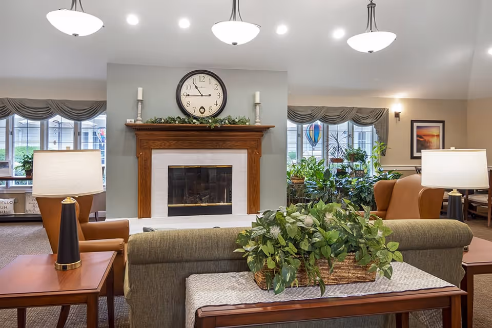 A cozy living room area with a central fireplace featuring a wooden mantle and a round clock above it. The room has two armchairs with side tables and lamps on either side, a sofa with a plant centerpiece on a table behind it, and large windows with valances letting in natural light. The space is decorated with various plants and framed artwork on the walls.