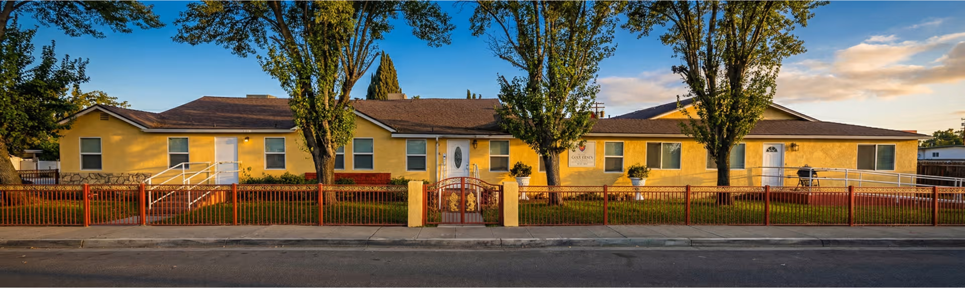 Single-story yellow senior living facility with a red fence and trees along the street.
