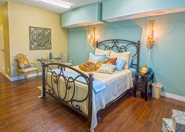 Bright, well-decorated bedroom with a wrought-iron bed, bedside tables and wall sconces against a teal accent wall, hardwood floors and a small seating area.