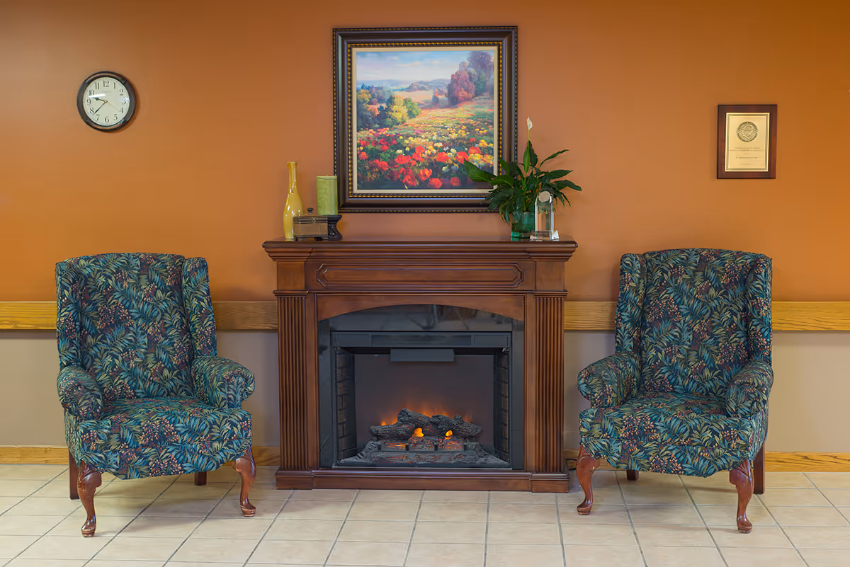Two floral patterned armchairs flank a wooden electric fireplace with a glowing fire. Above the fireplace hangs a colorful landscape painting of a field with flowers and trees. A clock showing 9:50 is mounted on the orange wall to the left, and a framed certificate is on the right. Decorative items including vases and a plant are placed on the fireplace mantel.
