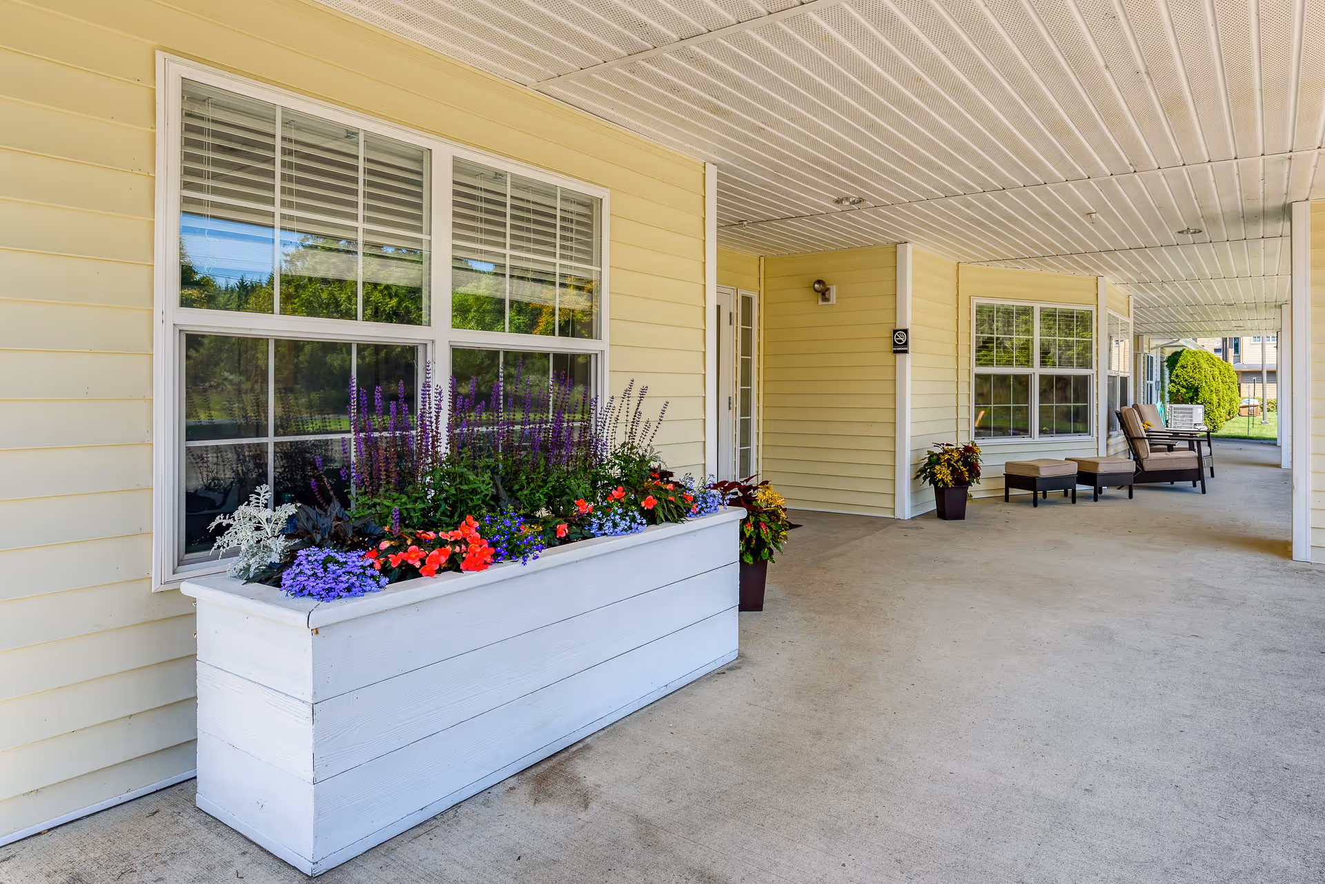 Covered entrance walkway with a large white planter of colorful flowers and outdoor seating by a pale yellow building.