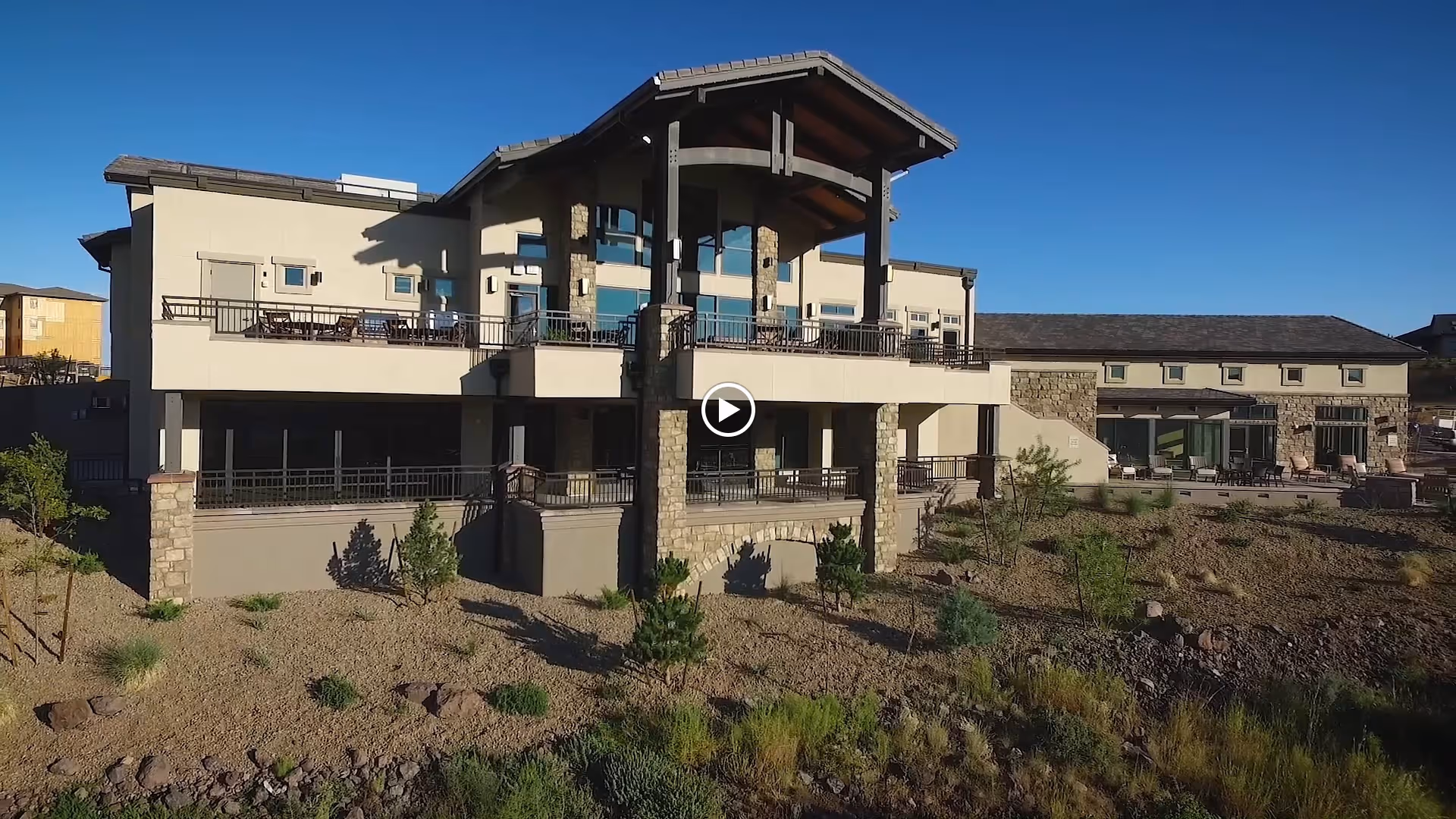 Exterior view of a large senior living facility building with stone and beige walls, multiple balconies, and a covered patio area, surrounded by landscaped dry terrain with small shrubs and plants under a clear blue sky.