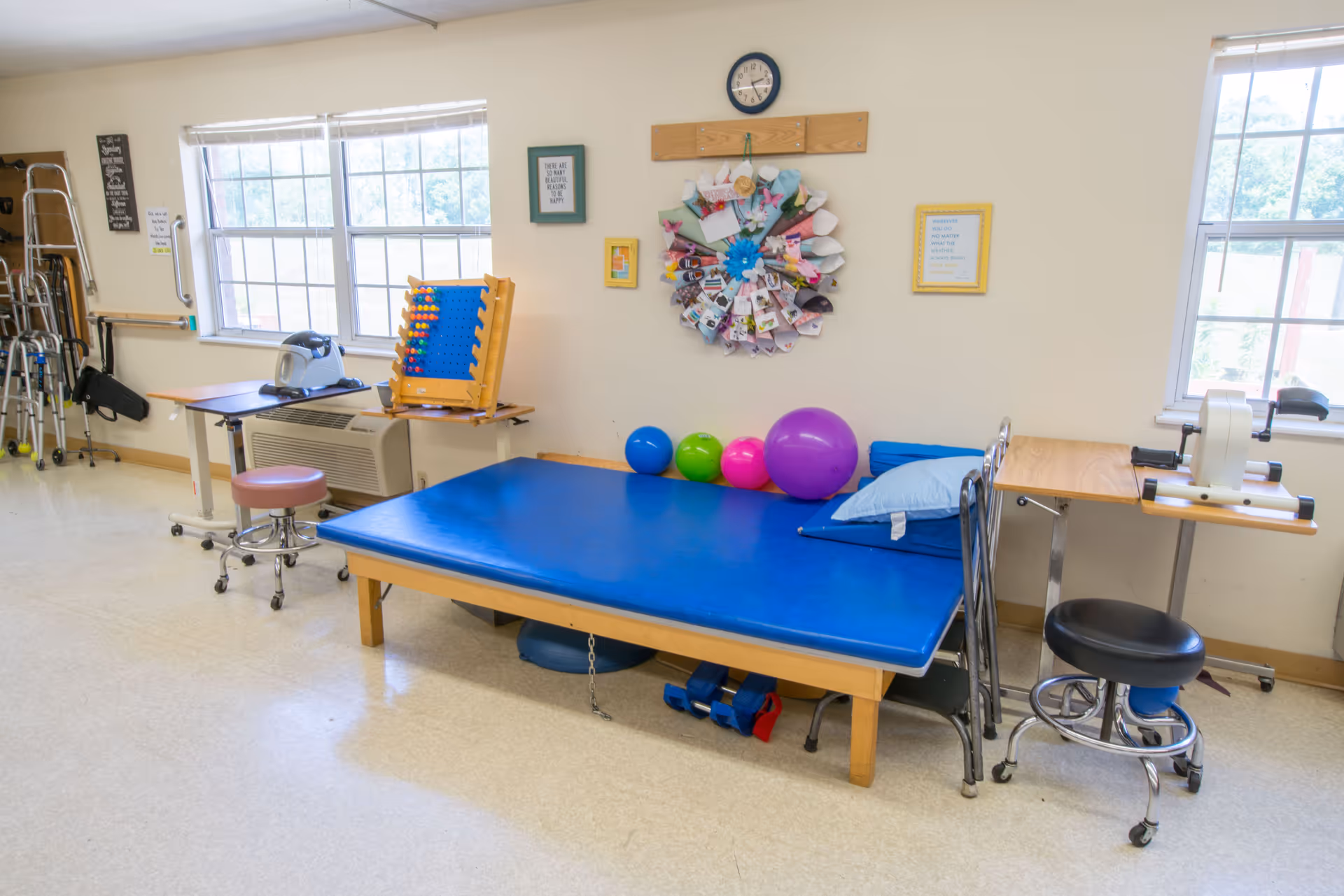 A physical therapy room with a blue padded therapy table, colorful exercise balls, a stool, and various therapy equipment including walkers and exercise devices. The room has two large windows letting in natural light and motivational wall decorations.