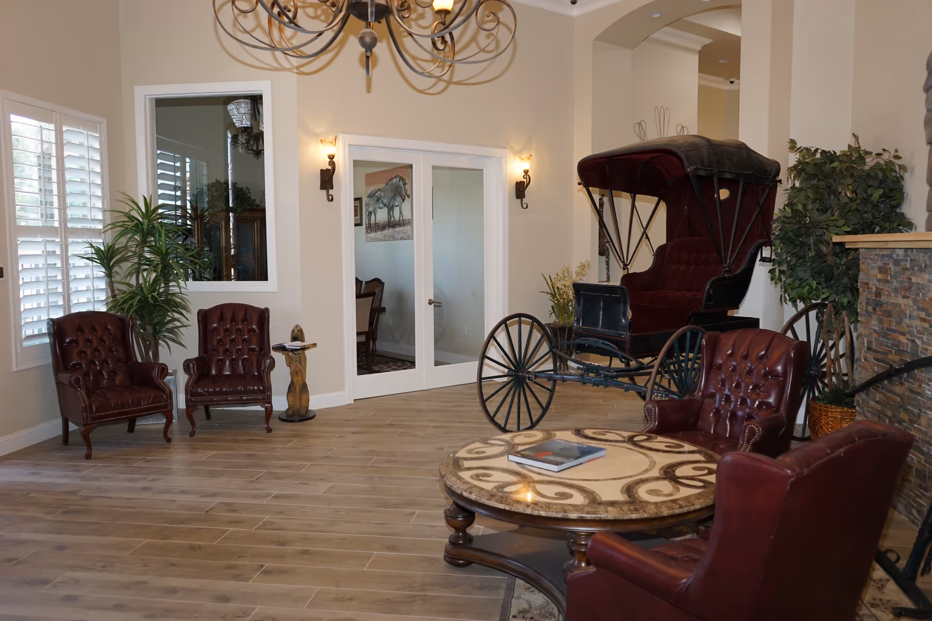 A cozy living room area with wooden flooring, featuring four dark red leather armchairs arranged around a round marble-top table with a book on it. There is a vintage horse-drawn carriage displayed in the corner, potted plants near the windows and fireplace, and double glass doors leading to another room. The walls are beige with wall sconces and a decorative chandelier hanging from the ceiling.