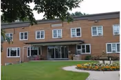 Brick senior living facility front entrance with glass doors, manicured lawn, and flower bed.