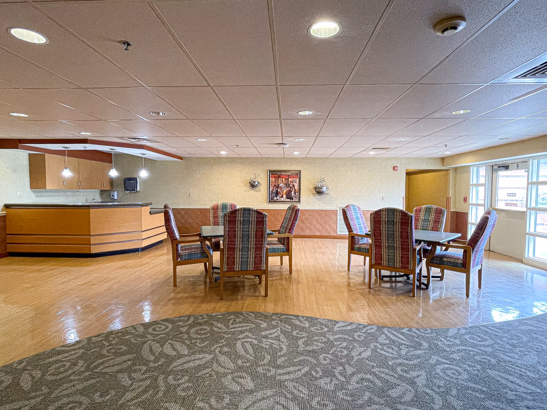 Spacious indoor senior living common area with dining tables, plaid chairs, and a front desk.