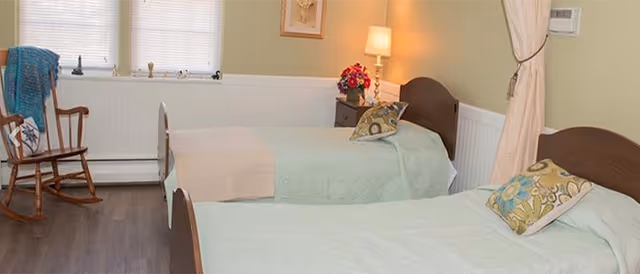 Two twin beds with pastel bedding, a nightstand lamp, and a rocking chair in a bedroom.