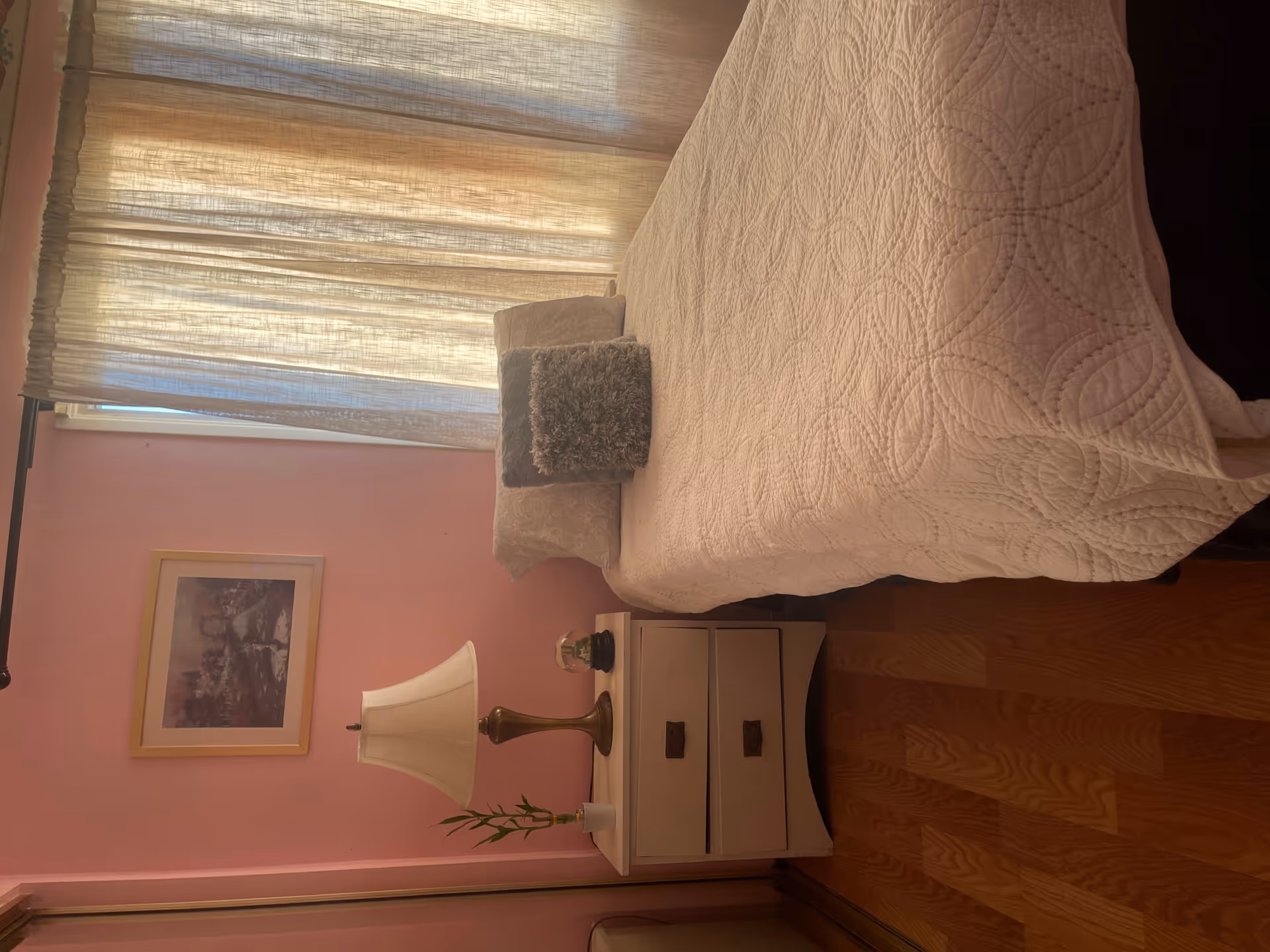 Pink bedroom with a quilted bed, nightstand, lamp, curtain window, and framed wall art.