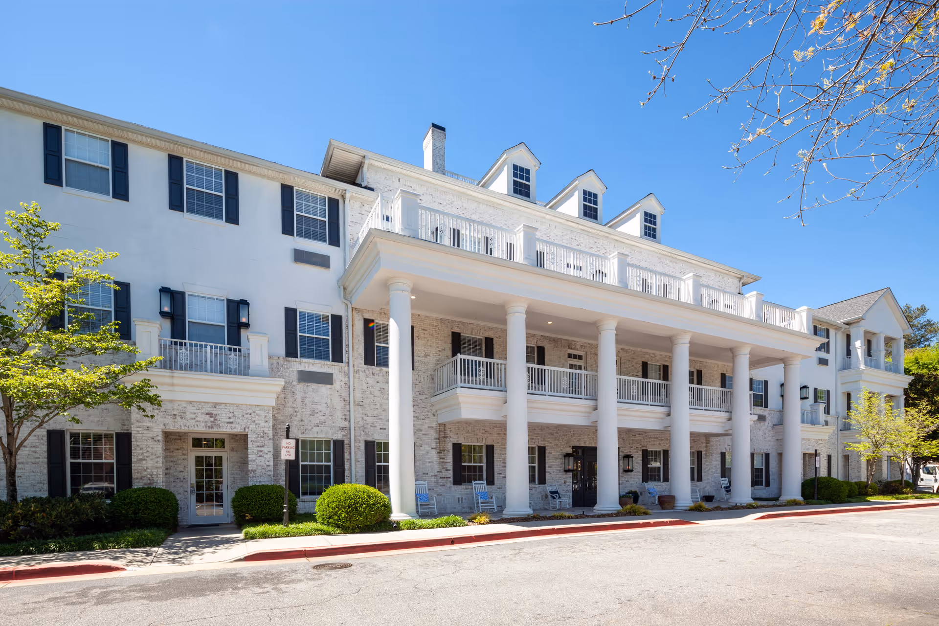Exterior front view of a large, three-story senior living facility building with white brick walls, black window shutters, and a covered porch supported by tall white columns. There are balconies on the second floor and a clear blue sky above.