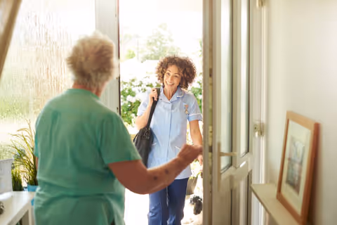 A smiling female healthcare worker in a light blue uniform enters a home, greeted by an elderly woman with short white hair wearing a green top. The scene is bright with natural light coming through the open door, and there is a framed picture on a small table inside the entrance.