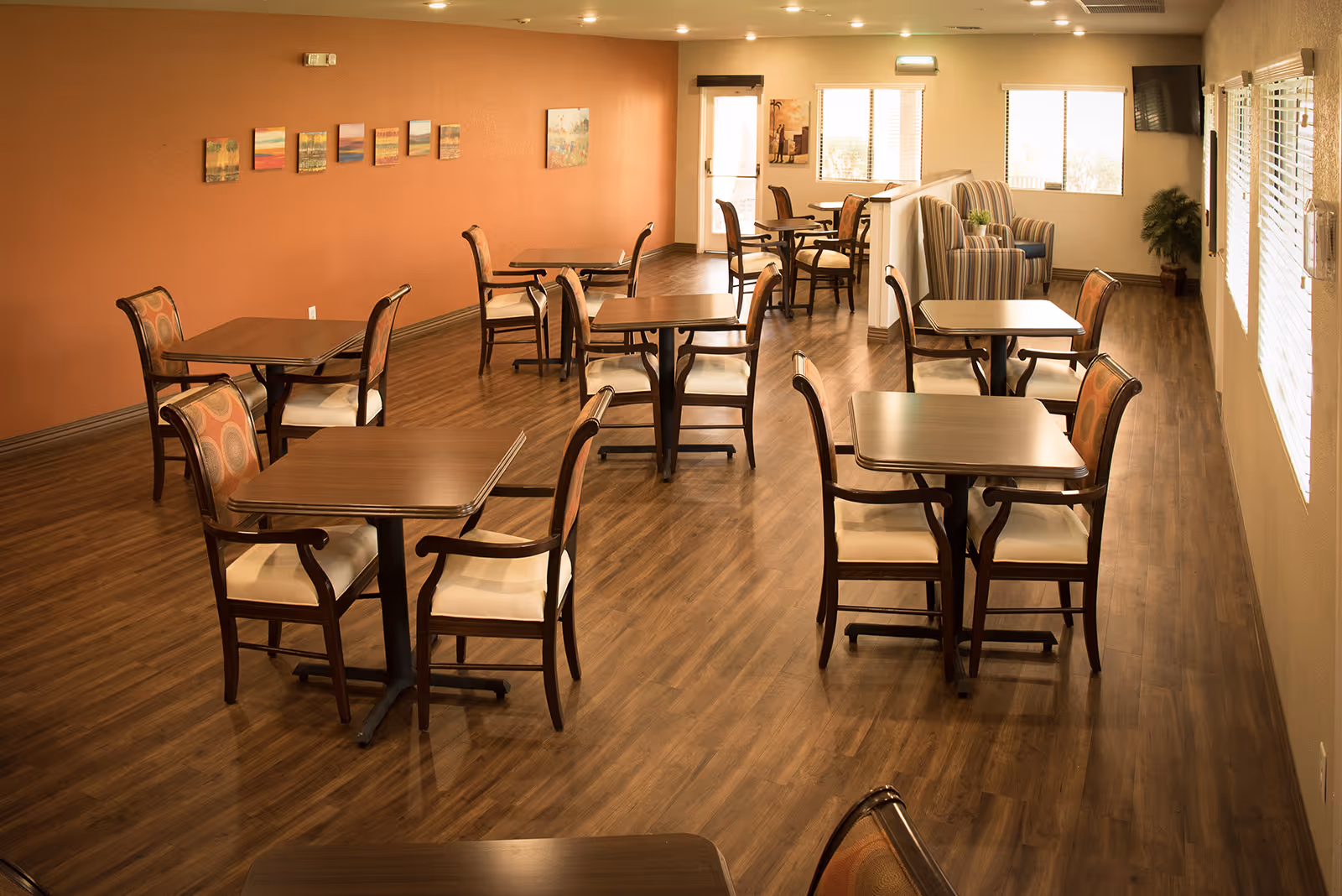 Well-lit communal dining room with multiple wooden tables and upholstered chairs on wood flooring and a seating area by the windows.