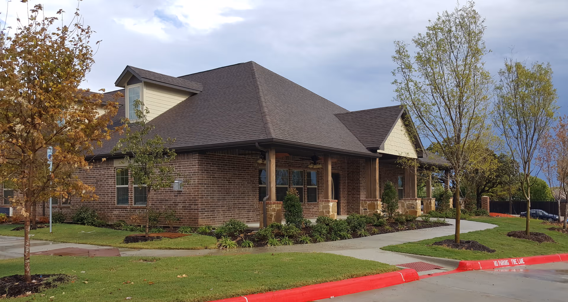 Exterior view of a single-story brick building with a dark shingled roof, surrounded by a well-maintained lawn, small trees, and shrubs. A sidewalk runs along the front of the building, and the sky is partly cloudy.