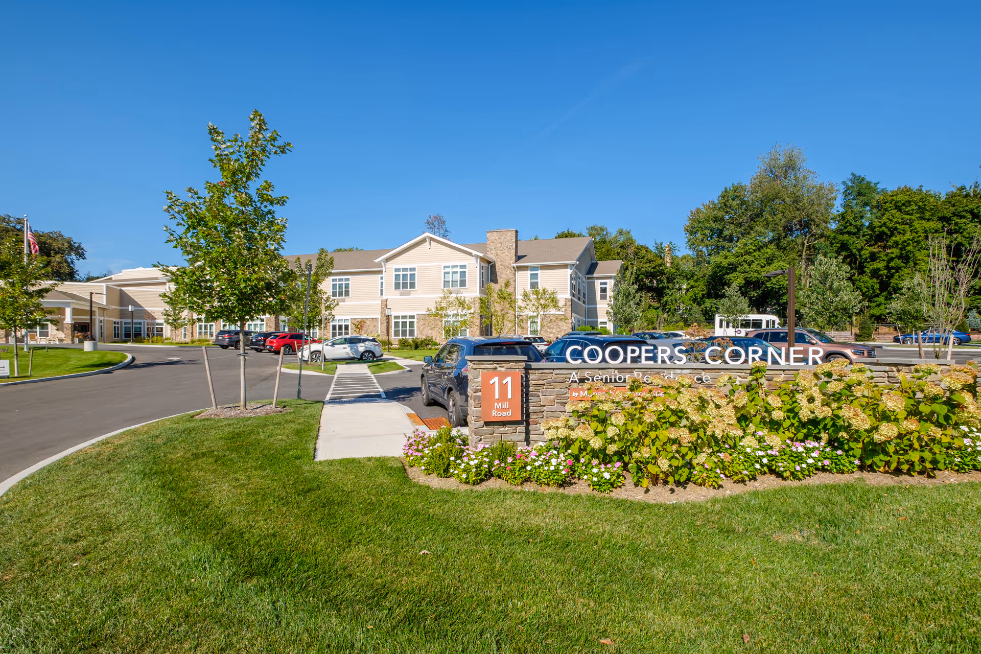 Front exterior of Coopers Corner senior living building with landscaped grounds, a stone sign, driveway and parked cars under a clear blue sky.