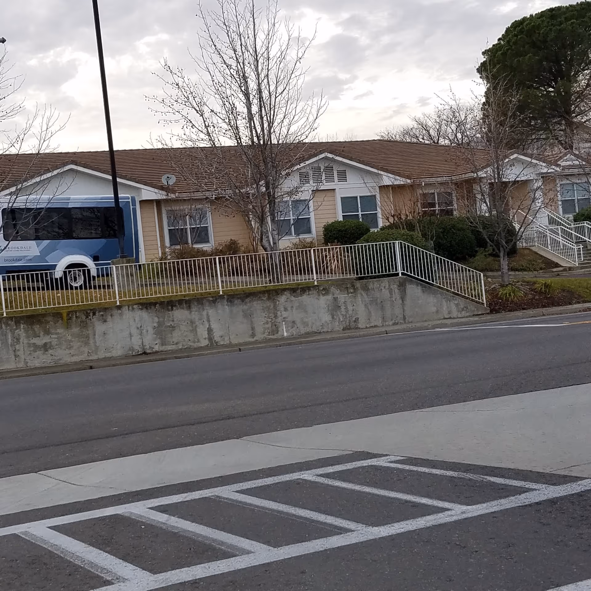 Single-story beige senior living facility along a street with a ramp, railings, and a blue bus.