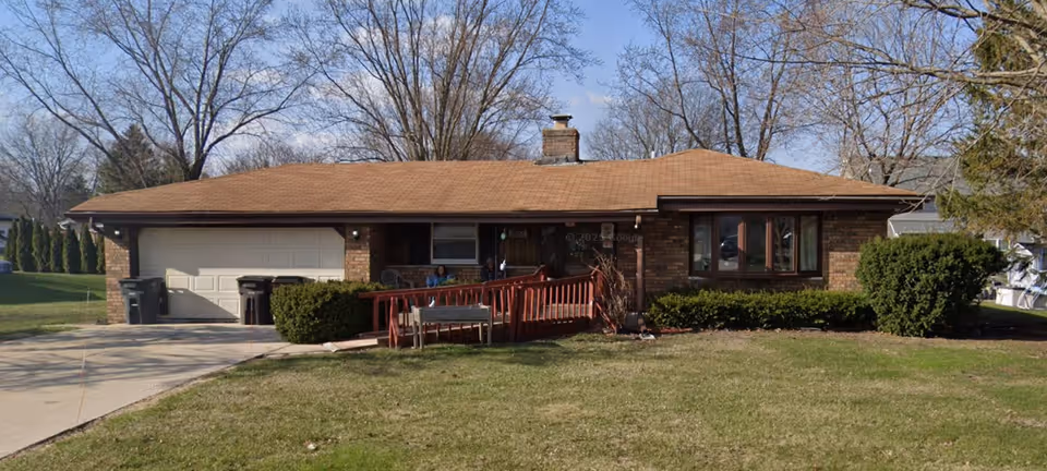 Single-story brick senior living facility exterior with ramp, driveway, and front yard.