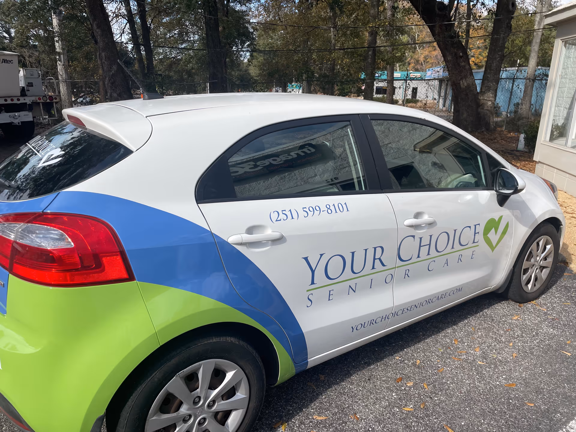 A white, blue, and green Your Choice Senior Care car parked in a lot beside trees.