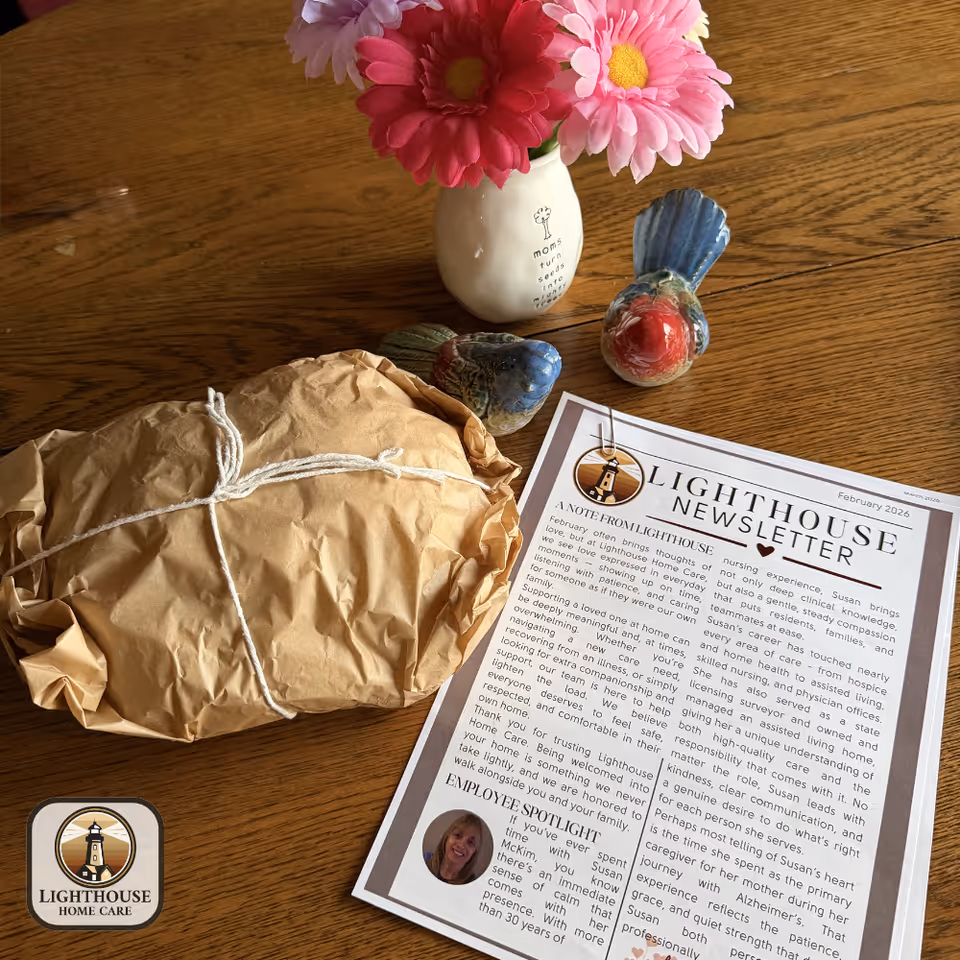 A wooden table holds pink flowers, a wrapped package, decorative birds, and a Lighthouse newsletter.