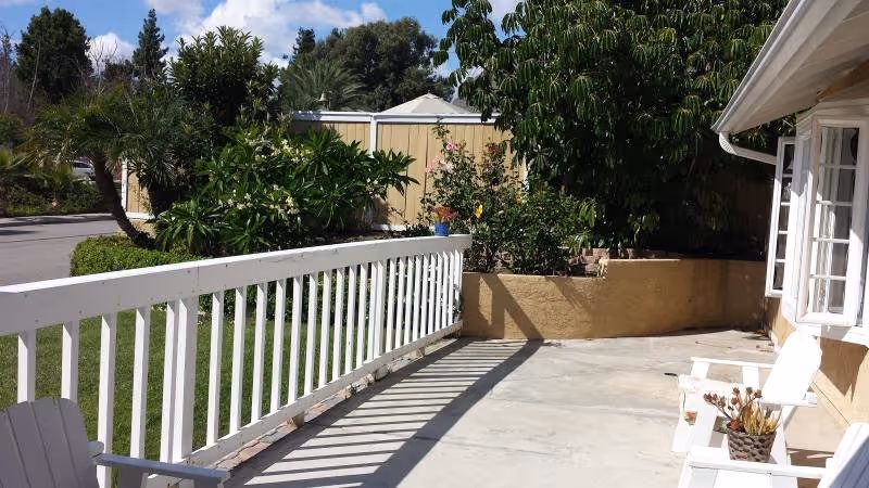 A sunny outdoor patio features a white railing, chairs, and potted plants beside a building.