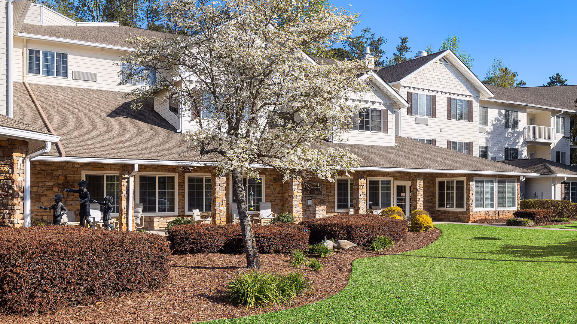 Exterior view of a senior living facility building with beige siding and stone accents, surrounded by well-maintained landscaping including a blooming tree, bushes, and green lawn under a clear blue sky.