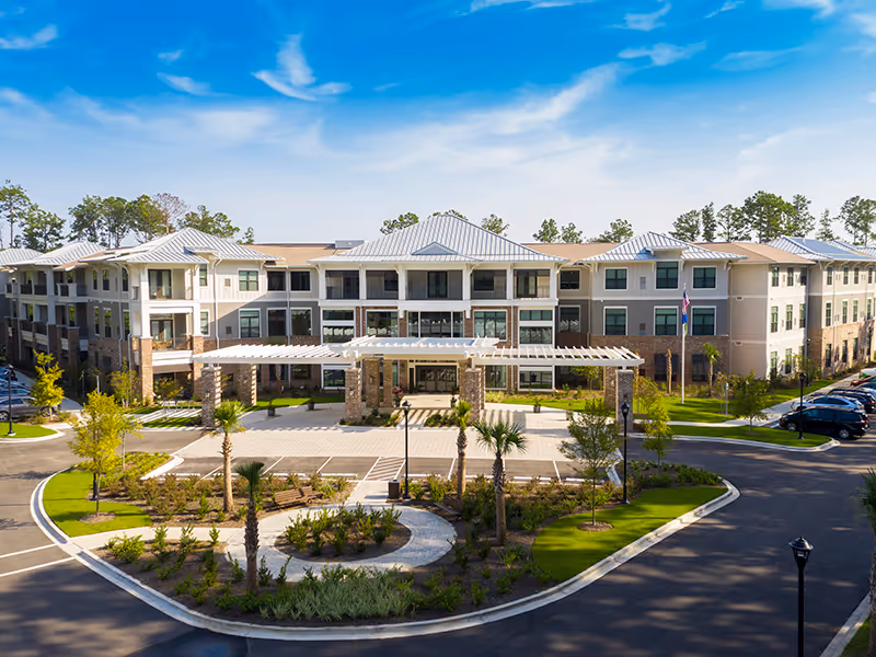 Front exterior view of The Artisan Mount Pleasant senior living facility, showing a large three-story building with a covered entrance, landscaped garden with palm trees, and a parking area under a clear blue sky.