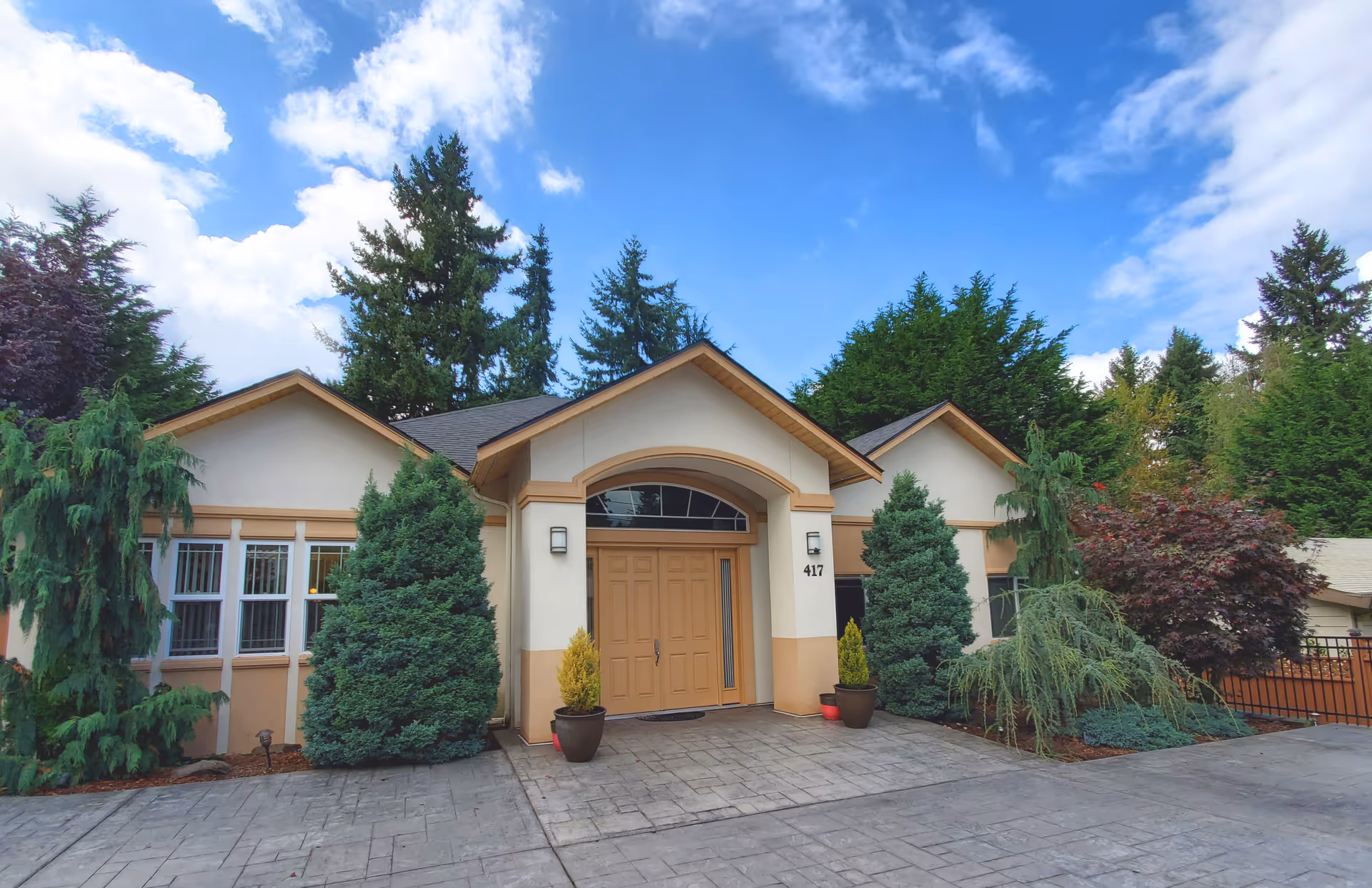 Front exterior of a single-story house-like building with a central double-door entrance, potted plants, and surrounding trees under a blue sky.