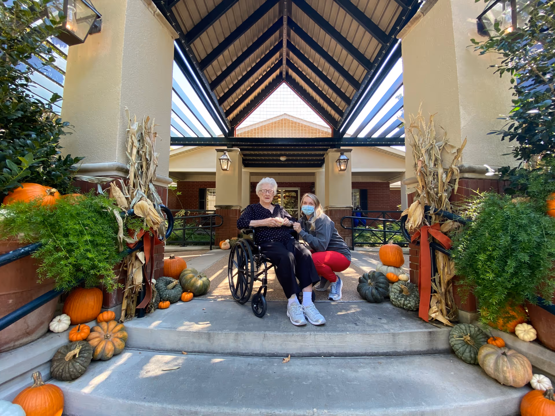 An elderly woman in a wheelchair and a younger woman wearing a face mask are posing together outside the entrance of a building. The entrance is decorated with various pumpkins, gourds, and dried corn stalks, suggesting a fall or Halloween theme. The building has a covered porch with a peaked roof and brick pillars.