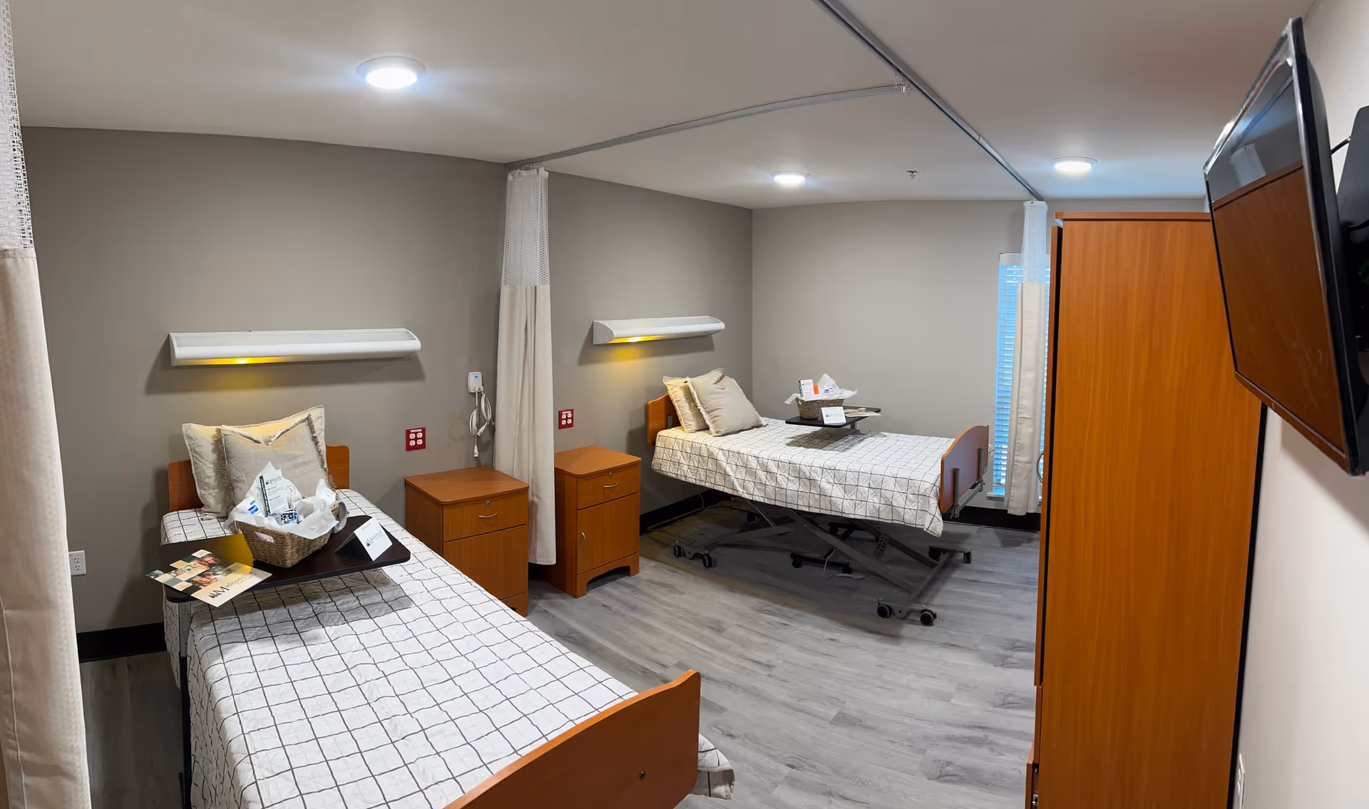 A clean and well-lit room with two single beds, each with a checkered bedspread and pillows. Between the beds are two wooden nightstands, and each bed has a wall-mounted light fixture above it. There is a wooden wardrobe on the right side of the room and a flat-screen TV mounted on the wall. The floor is covered with light-colored wood laminate, and the walls are painted a neutral gray. Privacy curtains hang between the beds and near the window.