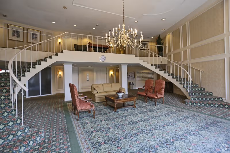 Spacious interior common area with a large patterned carpet, a beige sofa, four red upholstered chairs around a wooden coffee table, and a grand chandelier hanging from the ceiling. Two symmetrical staircases with white railings lead to an upper level with a railing. The walls are decorated with framed pictures and paneling.