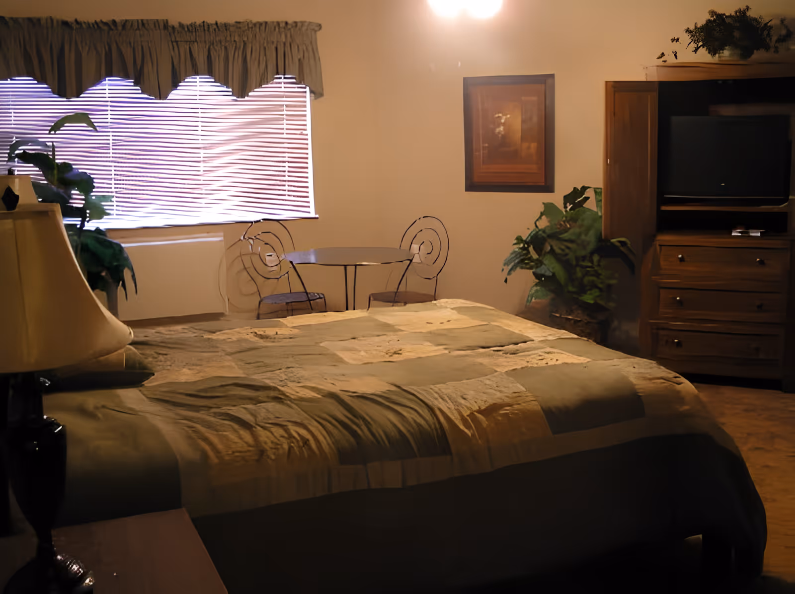 A warmly lit bedroom with a large bed, bedside lamp, dresser with TV, plants, and a small table and chairs by a window.