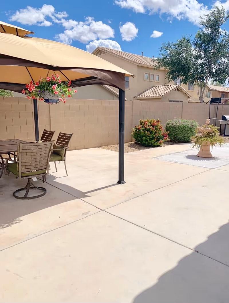 Outdoor patio area with a beige canopy providing shade over a table and four chairs. Hanging flower basket with pink and red flowers is attached to the canopy. In the background, there are bushes, a tree, and beige residential buildings under a partly cloudy blue sky.