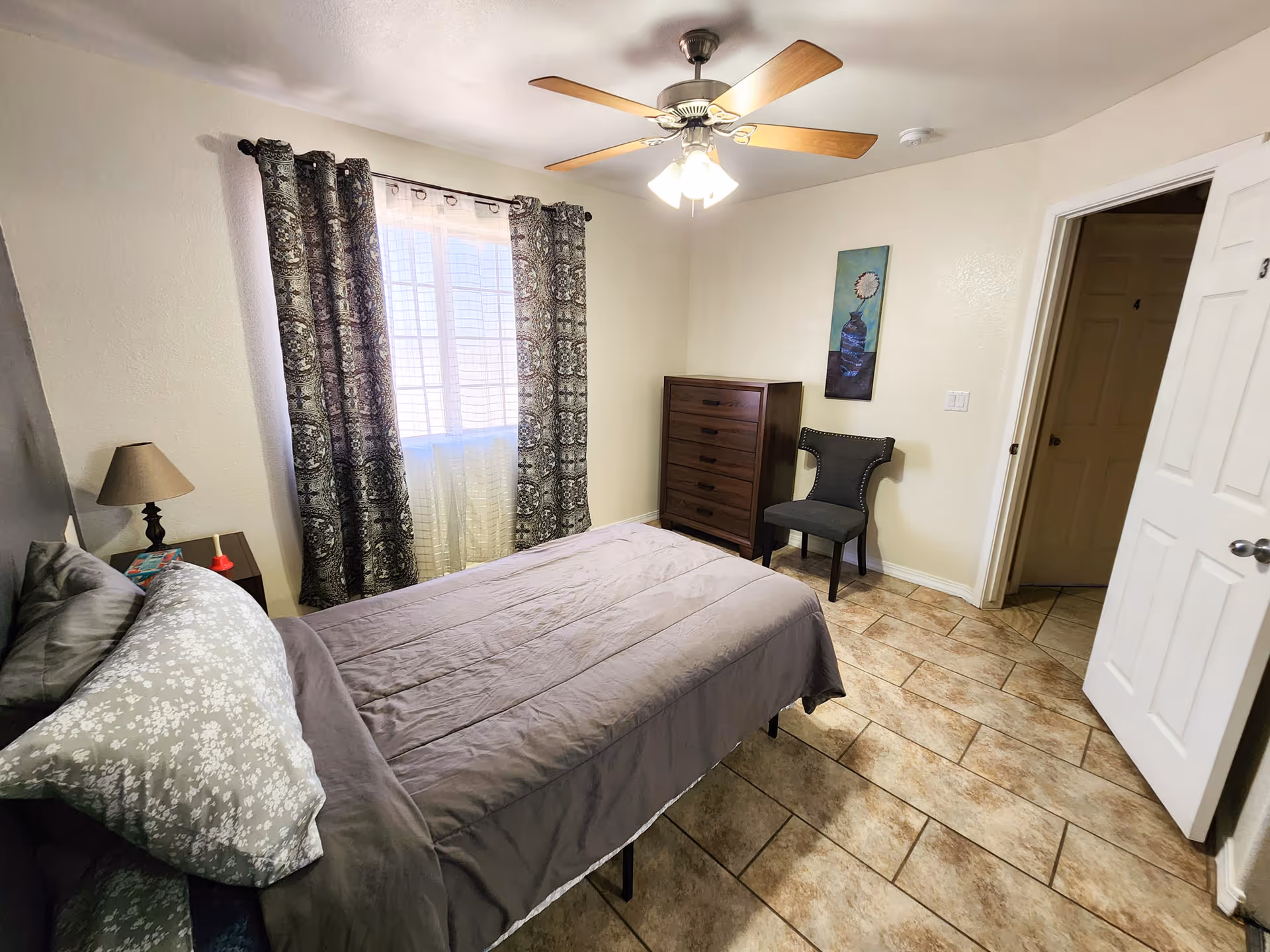 A furnished bedroom with a bed, dresser, chair, window with patterned curtains, and ceiling fan.