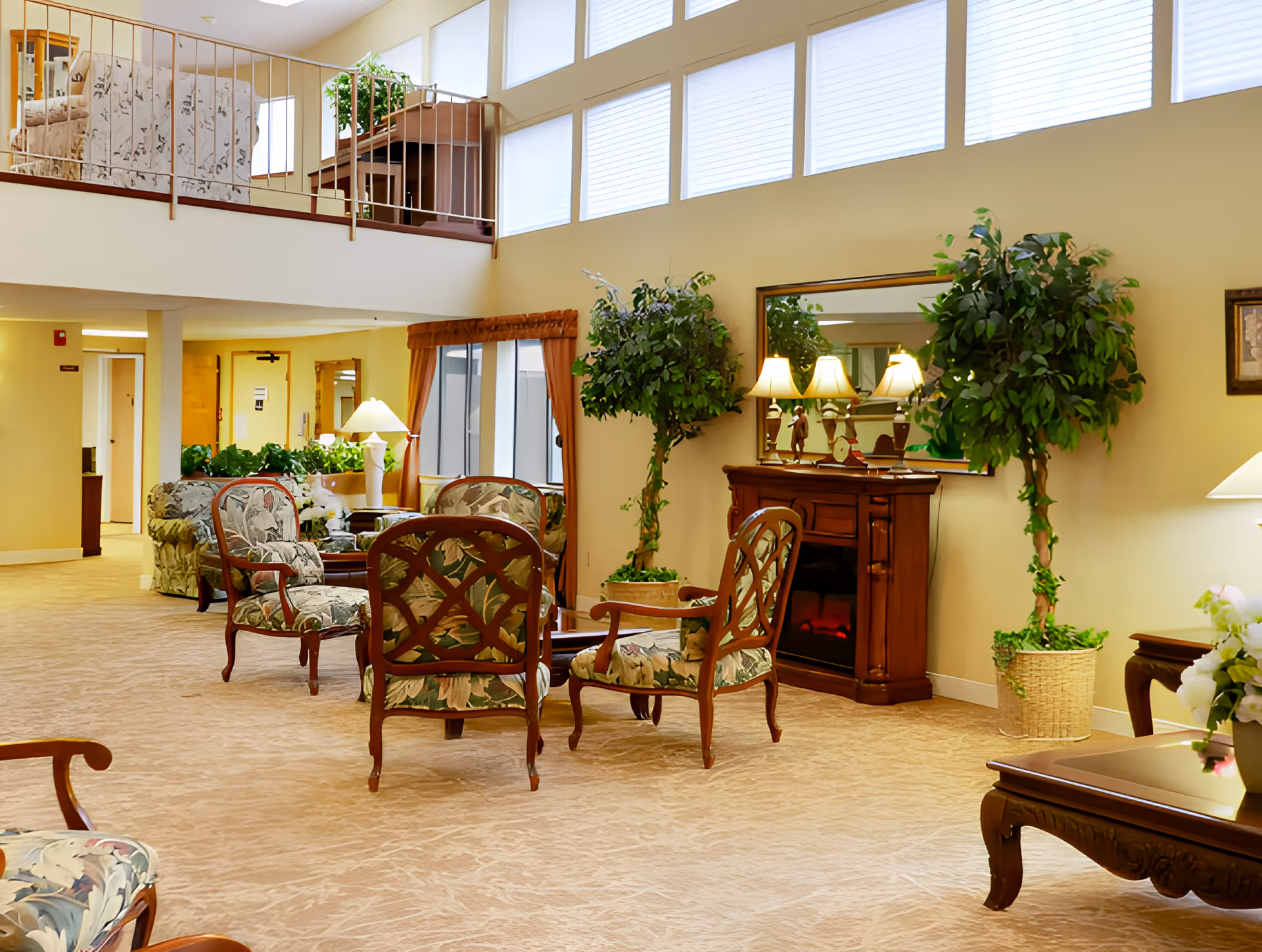 A spacious and well-lit senior living facility common area with floral upholstered chairs arranged around a wooden coffee table. The room features large windows with blinds, potted plants, a wooden electric fireplace with lamps on top, and a mirror above it. There is a staircase with a railing leading to an upper level with additional seating visible.
