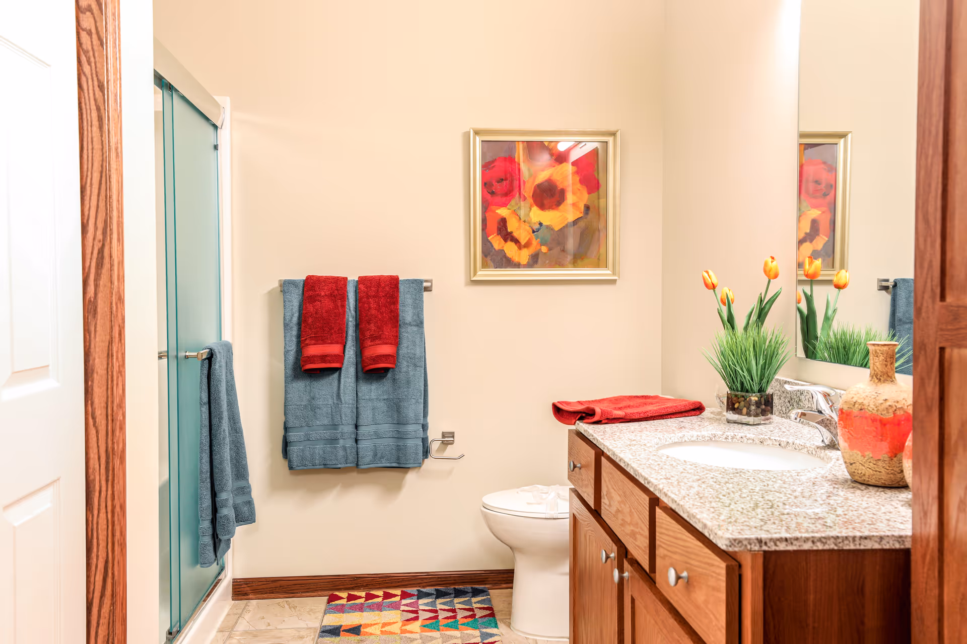 Bright bathroom with glass shower, sink, toilet, framed art, and blue-red towels.