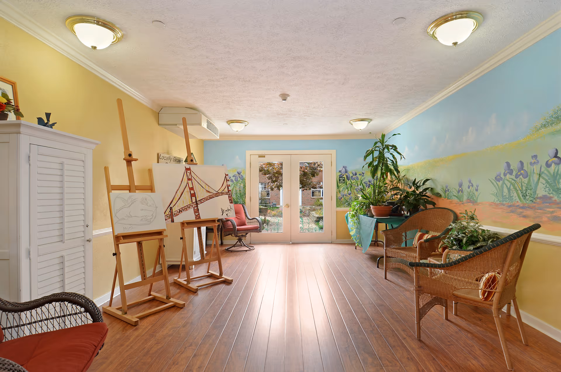 A bright, cozy room with wooden flooring and yellow and blue walls. The blue wall features a mural of flowers and a path. There are two wooden easels with paintings, wicker chairs with cushions, a small table with potted plants, and double glass doors leading outside.