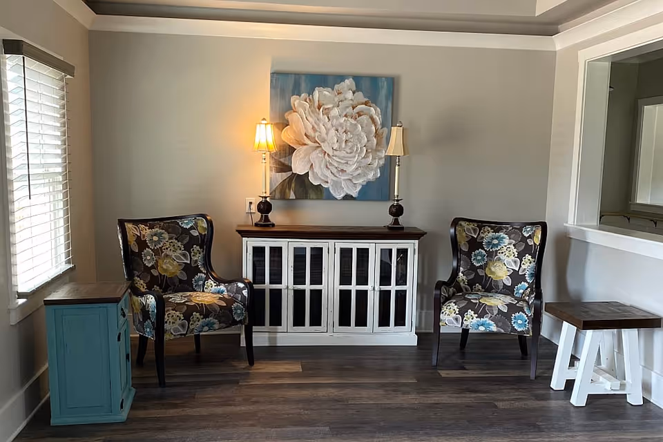 A cozy sitting area with two floral patterned armchairs placed on either side of a white wooden cabinet with glass-paneled doors. On top of the cabinet are two table lamps with beige shades and a large floral painting on the wall behind. To the left is a window with white blinds, and to the right is a small wooden table with white legs. The floor is dark wood, and the walls are painted light gray.